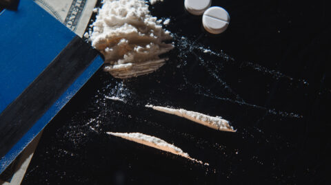 A close-up of white powder arranged in lines on a dark surface beside a stack of cash, two white pills, and a blue notebook, suggesting illicit drug use or possession.