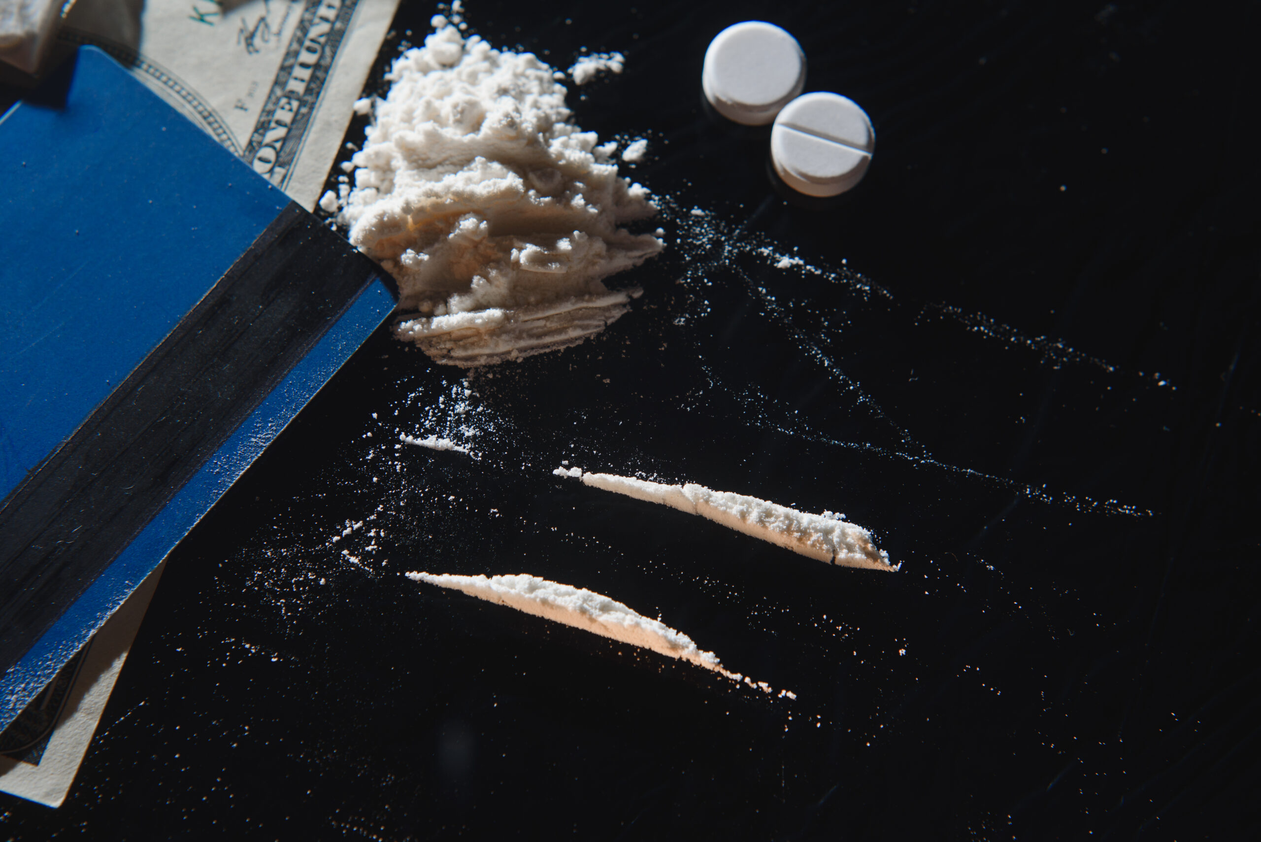 A close-up of white powder arranged in lines on a dark surface beside a stack of cash, two white pills, and a blue notebook, suggesting illicit drug use or possession.