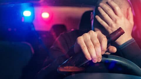 Driver holding beer with police lights in background image