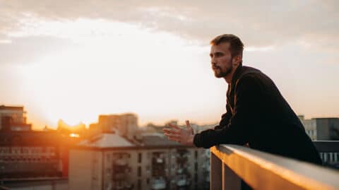 Man looking thoughtfully out at city from balcony during sunset