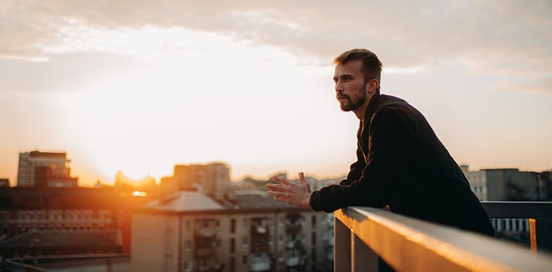 Man looking thoughtfully out at city from balcony during sunset