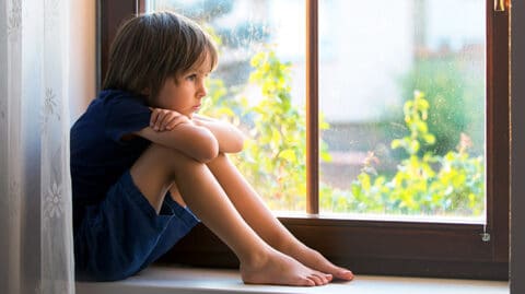 Young boy sitting alone on bench near window looking outside