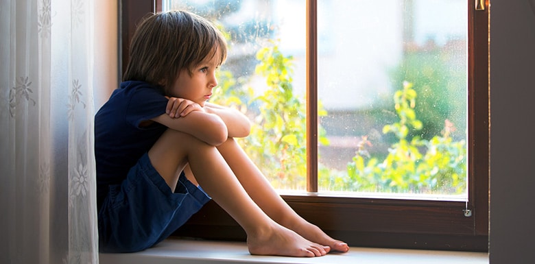 Young boy sitting alone on bench near window looking outside